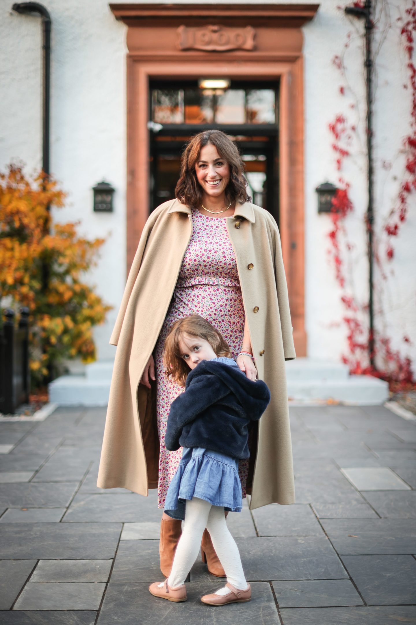 A woman and little girl standing together looking to camera in front of a hotel front door with autumn leaves in shot